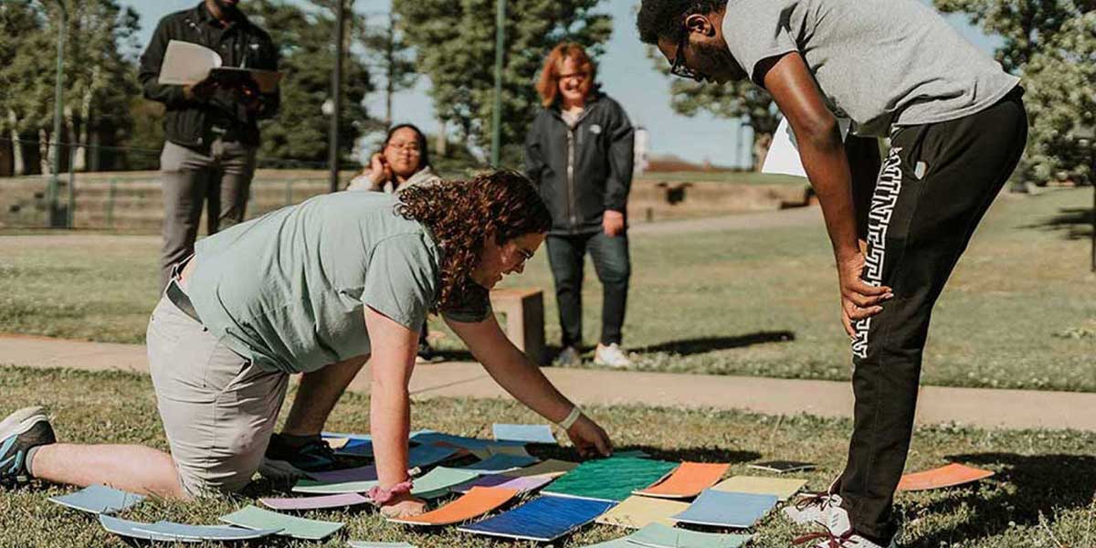 A student crouches down on the grass field to flip colorful cards as another student observes on the side